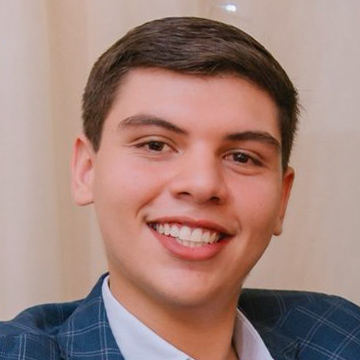 Nicolas, aluno de Engenharia Mecânica, posando em um ambiente interno com um fundo claro. Ele usa um blazer xadrez e tem cabelos escuros, com um sorriso largo.