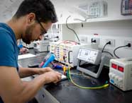 Aluno do curso de Engenharia Mecânica realiza atividade prática em laboratório de eletroeletrônica da Unoeste. Ele manuseia fios e instrumentos de medição sobre bancada preta, cercado por equipamentos como osciloscópio, multímetro digital azul e fonte de alimentação com display vermelho, demonstrando ambiente moderno e técnico.