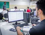 Estudante de Engenharia Mecânica trabalha em sala de informática da Unoeste, sentado diante de monitor com gráficos e códigos, caderno aberto, caneta e calculadora ao lado. Ao fundo, outros alunos utilizam computadores, enquanto um professor explica conteúdos escritos no quadro verde, compondo ambiente acadêmico focado em tecnologia.