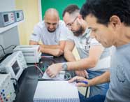 Grupo de estudantes de Engenharia Mecânica participa de atividade prática em laboratório da Unoeste. Eles analisam circuitos eletrônicos sobre bancada equipada com fontes, osciloscópios e anotações em papel. O ambiente é claro, com paredes claras e quadro verde ao fundo, ressaltando o caráter colaborativo e experimental do curso.