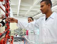 Alunos do curso de Engenharia Mecânica da Unoeste realizam experimento em laboratório de hidráulica. Vestindo jalecos brancos, manipulam tubos transparentes e vermelhos conectados a dispositivos de medição. O espaço possui teto branco quadriculado e equipamentos modernos, enfatizando o aprendizado prático em engenharia.