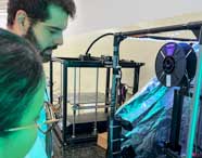 Dois estudantes observam atentamente uma impressora 3D em funcionamento no laboratório de Engenharia Mecânica da Unoeste. A máquina está envolta por estrutura metálica e lona refletiva azulada, com carretel de filamento preto visível. Ambos usam jaleco branco, destacando o ambiente inovador e tecnológico.
