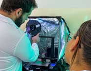 Aluno da Engenharia Mecânica da Unoeste, de jaleco branco, manipula carretel de filamento em impressora 3D dentro de gabinete revestido com material refletivo azul. Outra estudante observa atenta, ambos envolvidos em atividade prática de prototipagem rápida, ilustrando o uso de novas tecnologias no curso.