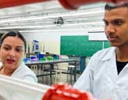 Laboratório de hidráulica da Unoeste: dois estudantes de Engenharia Mecânica em jaleco branco realizam experimentos com tubos vermelhos e transparentes. Ao fundo, quadro verde com inscrições "#AQUIÉUNOESTE" e "#MENINASNASTEM", mesas de trabalho e diversos equipamentos, mostrando ambiente colaborativo e moderno.