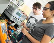 Dois estudantes de Engenharia Mecânica trabalham juntos em bancada de laboratório de eletrônica da Unoeste. Eles utilizam sondas e analisadores para medir componentes em placa de circuito, rodeados por instrumentos como osciloscópio e multímetro laranja, demonstrando integração entre teoria e prática.