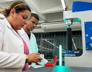 Estudante e professora de Engenharia Mecânica da Unoeste, ambos de jaleco branco, operam máquina de ensaio mecânico azul. A professora segura peça vermelha sobre a base do equipamento, enquanto o aluno observa. Ao fundo, laboratório amplo e bem iluminado, reforçando o caráter experimental do curso.