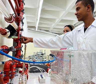 Estudante de Engenharia Mecânica, usando jaleco branco, manipula válvula em painel com tubos e conexões vermelhas, simulando sistemas hidráulicos. Na bancada, há equipamentos transparentes com água e instrumentos de medição, ambiente típico de laboratório universitário.