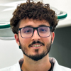 Imagem de Jean, aluno do curso de medicina no campus Guarujá, trajando jaleco branco. O fundo apresenta paredes claras, sugerindo um ambiente clínico ou laboratório técnico. A composição transmite seriedade e foco nos estudos médicos.