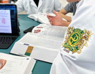 Estudantes de Medicina estudam juntos em mesa verde, utilizando livros abertos de anatomia e um tablet com imagem de coração. Em destaque, o brasão bordado do curso de Medicina da Unoeste Guarujá no jaleco branco, representando dedicação acadêmica e estudo colaborativo.