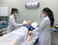 Duas alunas de Medicina da Unoeste Guarujá realizam atendimento simulado em manequim hospitalar adulto. Elas usam jaleco branco e protetor facial, monitorando sinais vitais em tela ao lado do leito, em ambiente de laboratório de simulação realística com equipamentos médicos.