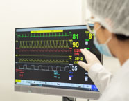 Aluno de Medicina da Unoeste Guarujá, com touca, máscara e luvas, aponta para monitor cardíaco digital. A tela exibe gráficos coloridos de frequência cardíaca, pressão arterial e saturação, simulando acompanhamento clínico detalhado em ambiente hospitalar de ensino.