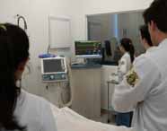 Alunos de Medicina em jalecos brancos observando um monitor médico que exibe gráficos de dados vitais de um manequim em um laboratório de simulação hospitalar.