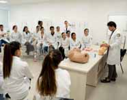 Professor de Medicina em jaleco branco realizando uma demonstração com um modelo de simulação de abdômen gestacional para um grupo de alunos atentos em sala de aula.
