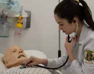 Aluna de Medicina em jaleco branco auscultando o tórax de um manequim de paciente com um estetoscópio, simulando um exame físico em ambiente hospitalar.