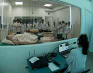 Vista de uma sala de controle com uma pessoa operando equipamentos, monitorando uma aula de simulação em tempo real, onde alunos de Medicina interagem com um manequim em laboratório.