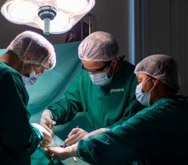 Três estudantes de Medicina da Unoeste realizam procedimento cirúrgico em centro cirúrgico, vestindo aventais verdes, toucas e luvas descartáveis. Uma luz cirúrgica intensa ilumina o campo operatório, enquanto os alunos manipulam instrumentos médicos com concentração e precisão.
