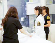 Três alunas de Medicina em jalecos brancos e scrubs escuros em laboratório de simulação, preparando manequim de paciente para procedimento, com monitor médico ao fundo.