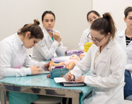 Grupo de alunas do curso de Medicina da Unoeste Jaú reunidas ao redor de uma mesa azul em sala de aula. Elas usam jaleco branco, escrevem em cadernos e interagem com um boneco infantil para treinamento, cercadas por estojos e materiais didáticos.