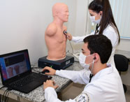 Dois estudantes de Medicina da Unoeste Jaú, de jaleco branco e estetoscópio, realizam exame clínico em manequim de simulação SAM conectado a um notebook. Um aluno opera o computador enquanto a colega ausculta o tórax do manequim em laboratório moderno.