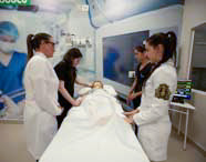 Quatro alunas de Medicina ou profissionais de saúde em jalecos e scrubs realizando simulação prática com manequim de paciente em ambiente hospitalar, com monitores ao fundo.