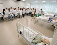Sala ampla da Unoeste Jaú com dezenas de estudantes de Medicina, todos de jaleco branco, assistindo aula teórica e prática. A professora ministra conteúdo ao lado de quadro branco e mesas, enquanto leitos hospitalares com manequins adultos e infantis ocupam o primeiro plano.