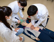 Três estudantes de Medicina da Unoeste Jaú, de jaleco branco, realizam simulação de primeiros socorros em manequim adulto no chão. Uma aluna faz ventilação com ambu, outra observa e um aluno executa massagem cardíaca, demonstrando treinamento em ressuscitação.