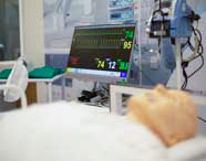 Monitor médico exibindo gráficos de batimentos cardíacos e dados vitais como FC, SpO2 e pressão arterial, com um manequim de paciente deitado em primeiro plano.
