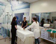 Três alunas de Medicina ou instrutoras em jalecos e scrubs, em laboratório de simulação hospitalar, observando e orientando junto a um manequim de paciente em uma cama.