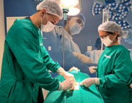 Estudantes de Medicina da Unoeste Jaú, vestidos com avental cirúrgico verde e touca, simulam procedimento cirúrgico em manequim sob luz cirúrgica intensa. O cenário reproduz ambiente de centro cirúrgico, com mural temático de profissionais em cirurgia ao fundo.