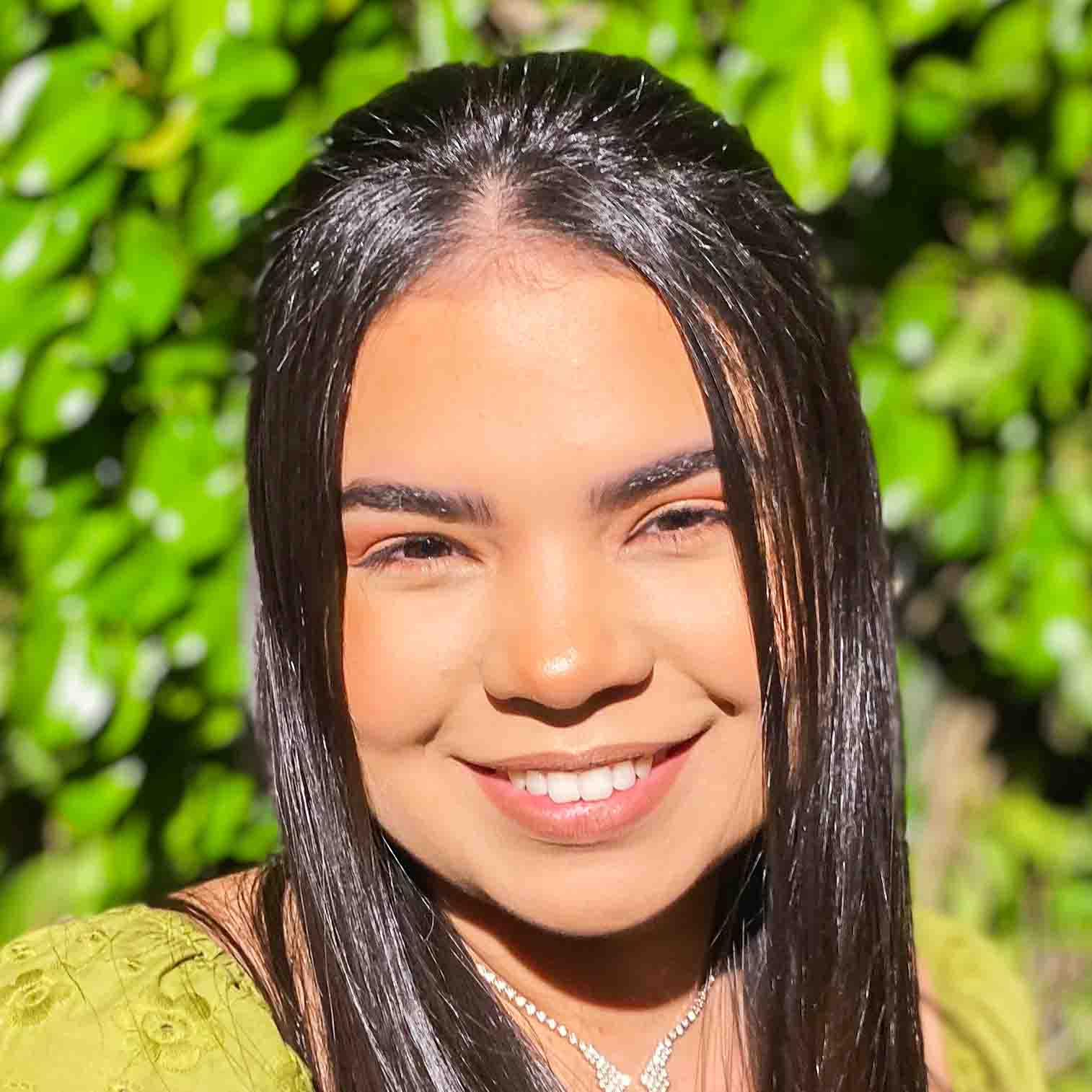 Alessandra, aluna do curso de farmácia, vestindo uma blusa verde com detalhes delicados. Ela está em um ambiente externo com fundo composto por folhagens verdes iluminadas pela luz natural, criando uma atmosfera vibrante e harmoniosa.