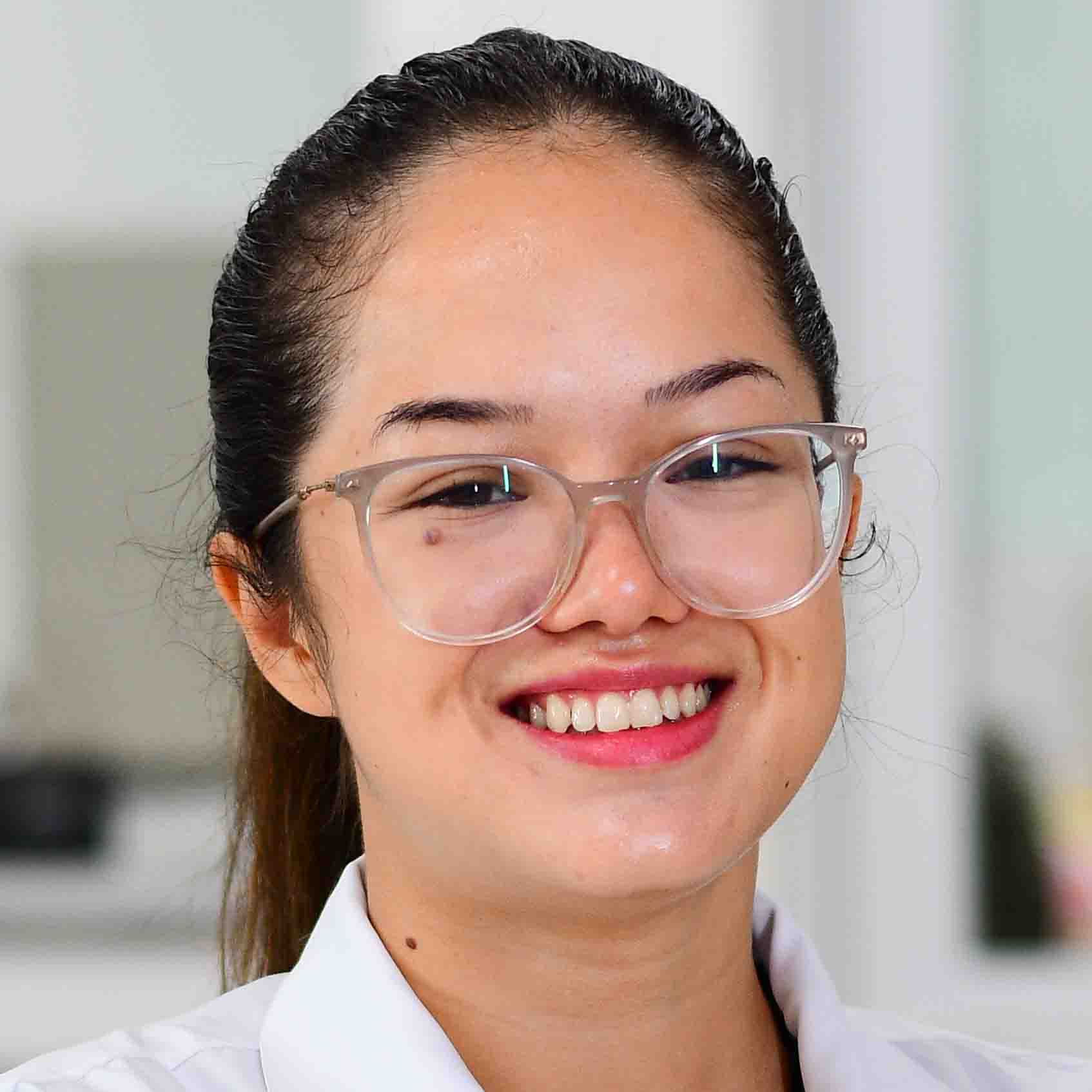 Bianca, aluna do curso de farmácia, trajando um jaleco branco. O ambiente ao fundo é interno, com tons claros e detalhes desfocados, sugerindo um espaço acadêmico ou laboratorial.