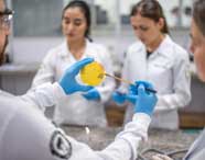 Estudantes do curso de Farmácia, usando jalecos brancos e luvas azuis, realizam atividade prática em laboratório. Um aluno segura uma placa de Petri amarela com cultura bacteriana, enquanto outro manipula a amostra com alça de inoculação. Ao fundo, colegas observam atentos ao experimento.