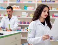 Dois estudantes de Farmácia, ambos de jaleco branco, atuam em ambiente de farmácia comunitária. Uma aluna lê atentamente uma receita, enquanto o colega organiza caixas de medicamentos nas prateleiras repletas de produtos farmacêuticos ao fundo.