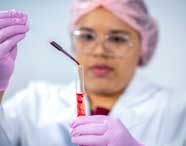 Pessoa com touca rosa, jaleco branco e luvas rosas manipula uma pipeta contendo sangue, transferindo o líquido para um tubo de ensaio. A imagem retrata atividade prática de análises clínicas no curso de Farmácia.