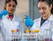 Duas estudantes de Farmácia, com jalecos brancos e luvas azuis, analisam tubos de ensaio com sangue em laboratório. Uma segura o tubo, enquanto a outra utiliza pipeta, cercadas por rack com outros tubos para exames clínicos.