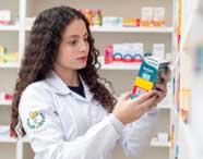 Aluna de Farmácia examina cuidadosamente uma caixa de medicamento na prateleira da farmácia comunitária da Unoeste. Ela veste jaleco branco com símbolo do curso bordado na manga, rodeada por estantes cheias de remédios organizados.