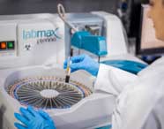 Estudante de Farmácia, com jaleco branco e luvas azuis, opera equipamento automatizado Labmax Plenno para análise clínica. Segura tubo de ensaio com amostra sanguínea, demonstrando prática laboratorial em ambiente moderno.
