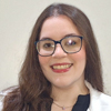 Ana, aluna do curso de biomedicina no campus Presidente Prudente, trajando jaleco branco. Ela está em um ambiente interno com fundo claro, sugerindo uma sala de aula ou laboratório acadêmico. A iluminação destaca o profissionalismo e o contexto educacional da imagem.