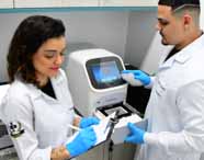 Biomedicina alunos em laboratório, dois estudantes com jalecos brancos e touca, luvas azuis, analisando resultados em um computador de bancada. O ambiente possui equipamentos laboratoriais, caixas organizadoras e iluminação fria, transmitindo ambiente de precisão e tecnologia.