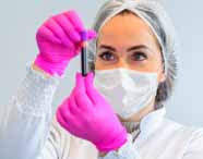 Análise de amostra de sangue, mulher de olhos claros usa touca, máscara e luvas rosa enquanto segura um tubo de sangue com tampa roxa, focando nos detalhes do exame. Fundo branco, uniforme branco com detalhes verdes, expressa cuidado e atenção no procedimento laboratorial.
