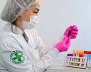 exames laboratoriais, estudante de jaleco branco, touca, máscara e luvas rosa observa um tubo de sangue. À sua frente há racks organizados com tubos de tampas coloridas, compondo um cenário limpo e organizado, típico de laboratório de análises clínicas.