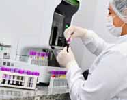 Análises clínicas, profissional com jaleco, touca e máscara realiza exame em tubo de sangue com tampa roxa. Diversos tubos organizados sobre bancada branca e equipamento automatizado ao fundo. O cenário destaca o ambiente clínico e a precisão do trabalho biomédico.
