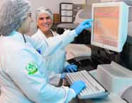 Estudantes com jaleco branco, touca e luvas azuis trabalham lado a lado em um equipamento de análises clínicas. Uma delas aponta para a tela de um monitor com interface laboratorial, enquanto a outra sorri. O cenário é um laboratório organizado, com luz suave e ambiente limpo.