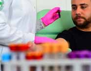 Estudante de biomedicina com jaleco branco e luvas rosas coleta sangue de um paciente sentado em uma cadeira verde. Em primeiro plano, tubos de ensaio coloridos desfocados compõem a cena. O paciente, com barba e camiseta preta, observa o procedimento atentamente.