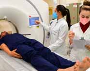  Profissionais de biomedicina acompanham um exame de tomografia computadorizada. Uma delas utiliza a tela do equipamento, enquanto a outra faz anotações em uma prancheta. Ambas usam jaleco branco e máscaras coloridas. Um paciente deitado usa roupa azul-escuro e entra no aparelho circular de tomografia.