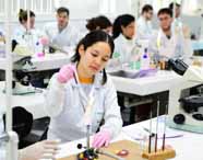 Aluna de biomedicina executa experimento de microbiologia, usando luvas rosas e jaleco branco. Ela segura uma alça de platina próxima à chama de um bico de Bunsen sobre uma bancada clara. No fundo, microscópios, tubos de ensaio e colegas em outras mesas de laboratório completam o ambiente.