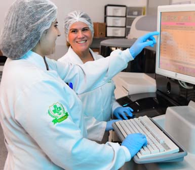  Duas mulheres em jaleco branco, touca e luvas azuis trabalham em laboratório. Uma aponta para monitor com tela alaranjada, a outra sorri diretamente para a câmera. Ao fundo, caixas organizadoras e equipamentos de laboratório em tons neutros completam o ambiente clínico e moderno.