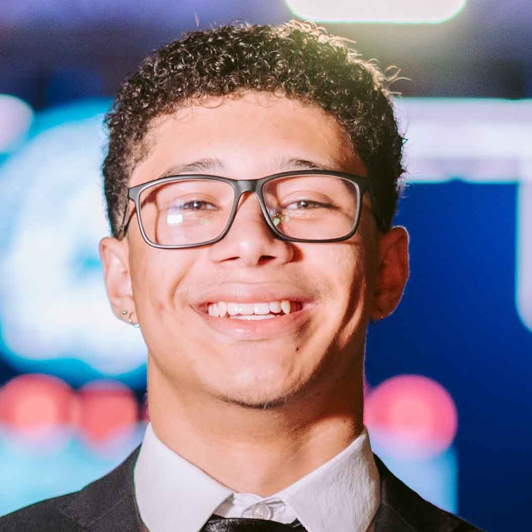 João Pedro, aluno de Engenharia de Produção, posando em um ambiente interno com luzes desfocadas ao fundo. Ele usa óculos de armação preta e um terno, com um sorriso largo.