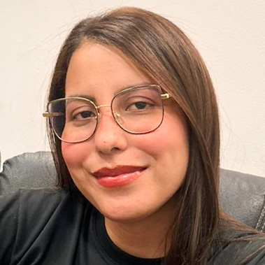 Luana, egressa de Engenharia de Produção, posando em um ambiente interno com um fundo claro. Ela usa óculos de armação dourada e tem cabelos castanhos, com um sorriso suave.