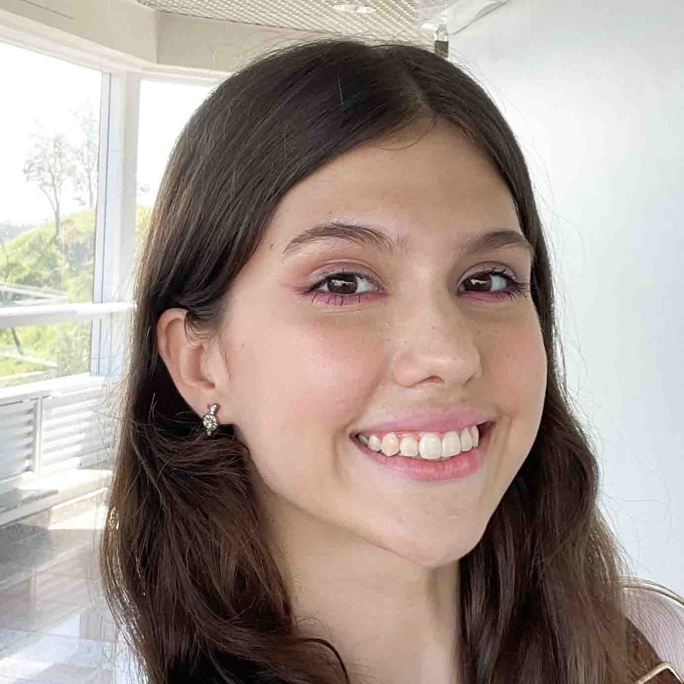 Maria Carolina, aluna de Engenharia de Produção, posando em um ambiente interno com uma janela ao fundo. Ela tem cabelos castanhos e um sorriso largo.